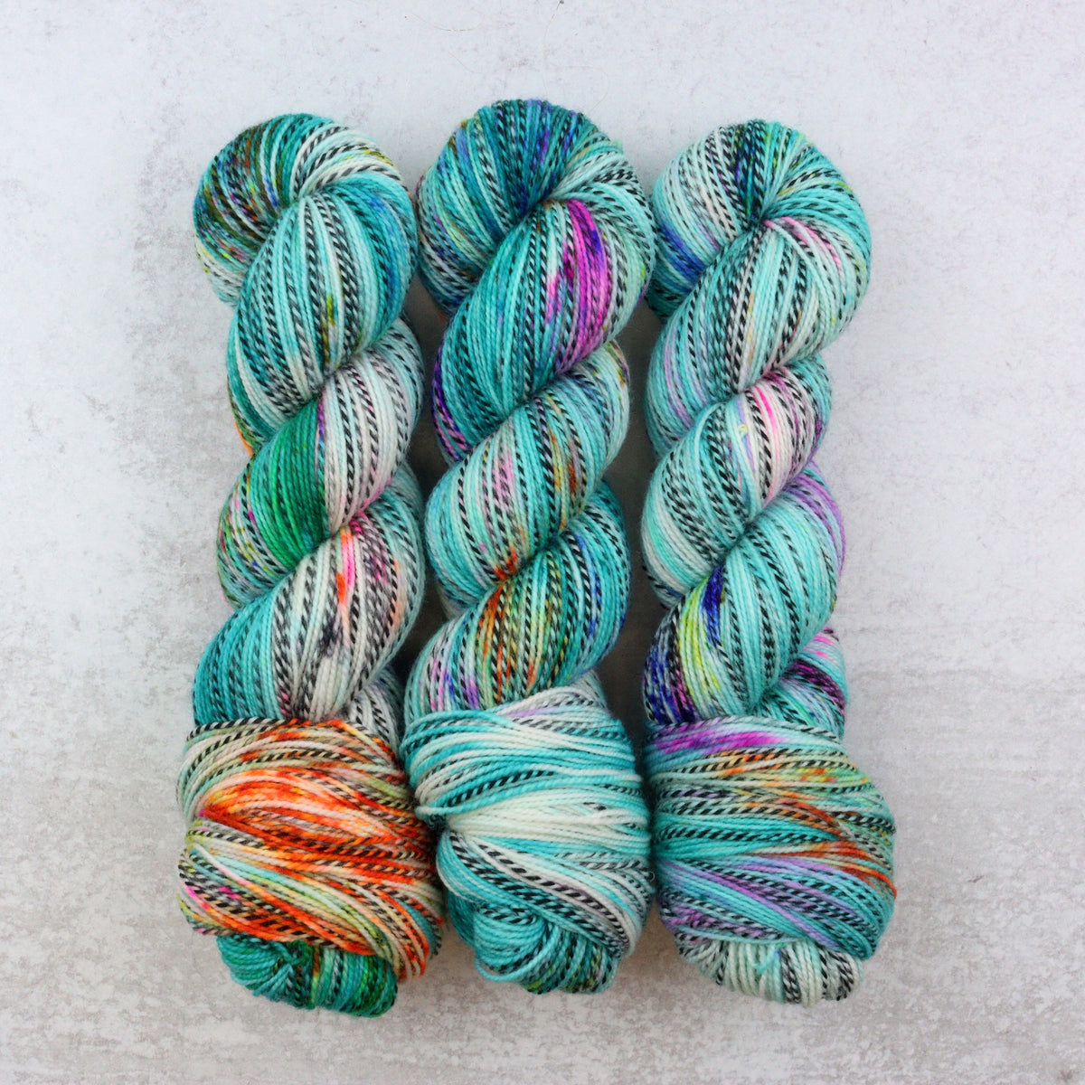 Three hanks of variegated yarn in shades of teal, mint, green, purple, orange, and white, arranged side by side on a light gray textured background.