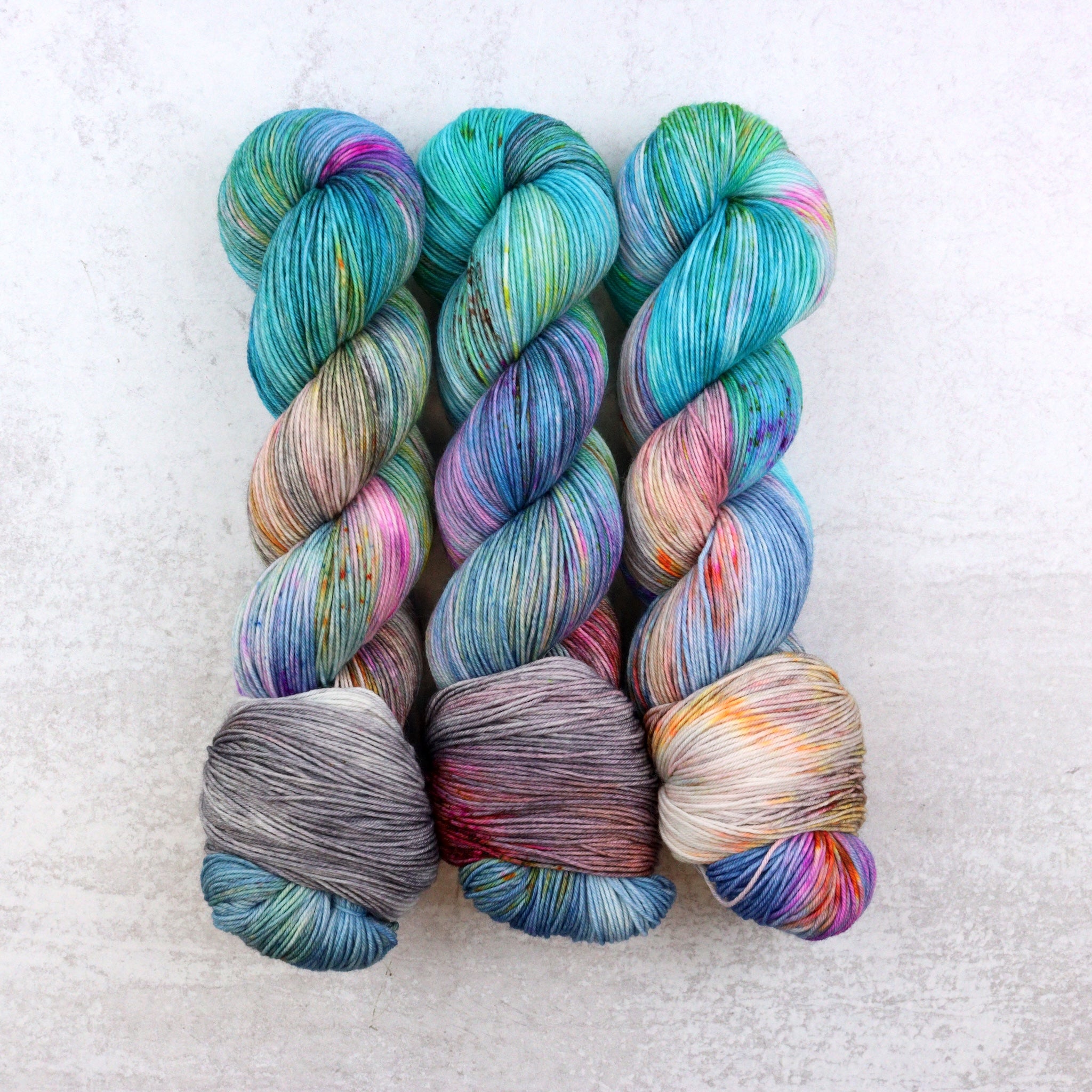 Variegated hand dyed sock yarn by Spun Right Round. Features pink, aqua, orange, green and brown speckles on a grey and aqua base.