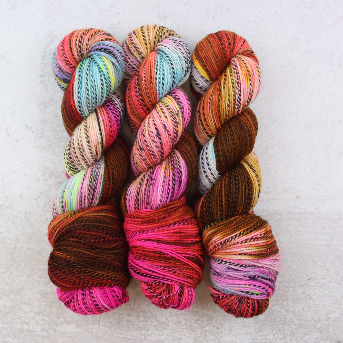 Three skeins of variegated yarn arranged vertically on a light textured background. The yarn features multiple colors including pink, orange, brown, blue, yellow, and white in a twisted variegated pattern.