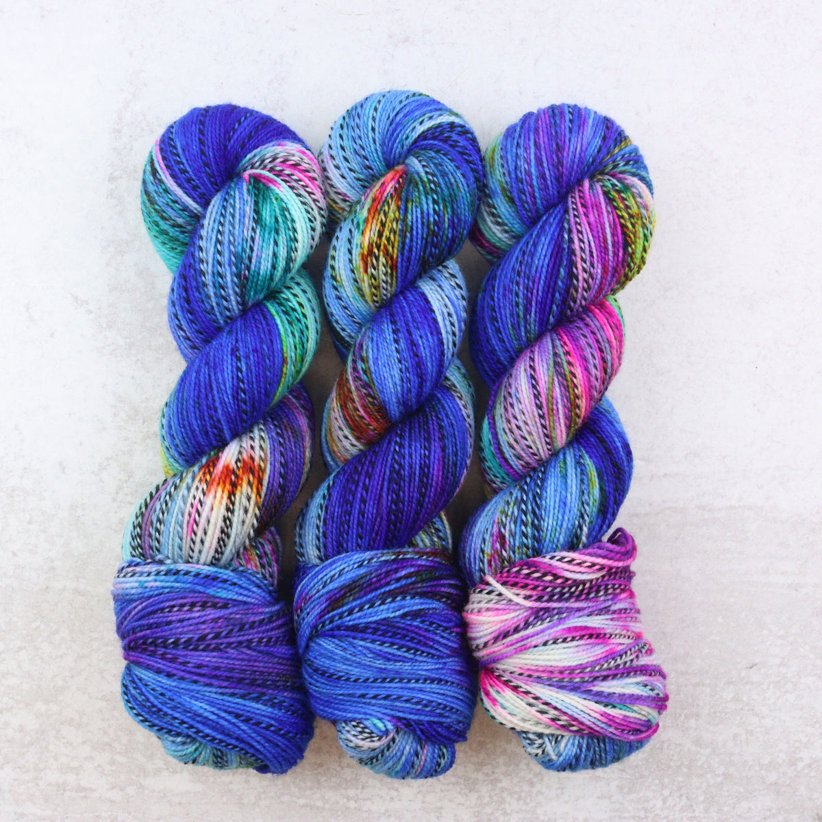 Three skeins of variegated yarn arranged vertically on a light textured background. The yarn features a multicolor variegated pattern with prominent shades of blue, purple, teal, pink, orange, and white.