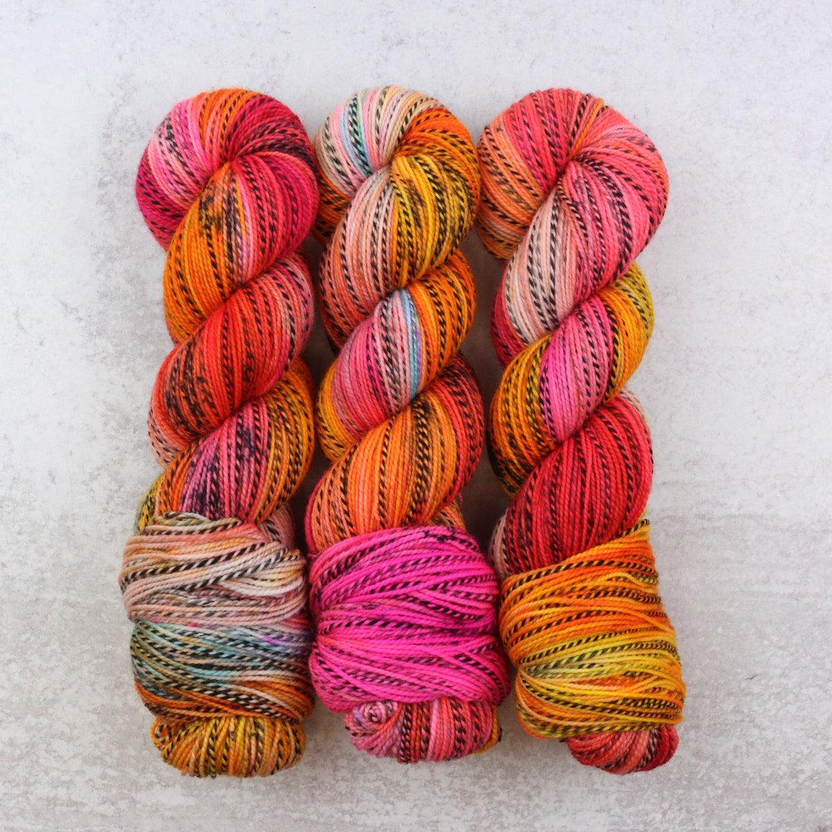 Three twisted skeins of variegated yarn arranged side by side on a light gray textured background. The yarns display a rich mix of colors including orange, pink, red, yellow, blue, and black, showcasing the variegated pattern.